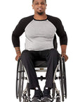 A man with a beard sitting in a manual wheelchair, wearing a black and gray raglan t-shirt, black pants, and black sneakers, photographed against a solid white background.