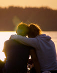 Two people sitting together with a dog at sunset by a body of water.
