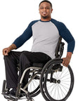 A smiling man in a gray and navy raglan long-sleeve shirt and black pants sits in a manual wheelchair against a plain white background.