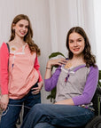 One woman standing in Sunset Raglan Colorblock Access tee, one woman in wheelchair in purple Raglan Access Tee, both showing chest port access