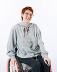Woman in wheelchair wearing gray zipper access hoodie with open chest zipper with port showing. Text reads "Katelyn is 5'6" wearing a size M".