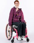A smiling person with short red hair and glasses sits in a pink manual wheelchair, wearing Plum hoodie with zippers open, black pants, and white sneakers against a plain white studio background. Small text at the bottom reads, 'Katelyn is 5'6" wearing a size M'.