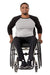 A man with a beard sitting in a manual wheelchair, wearing a black and gray raglan t-shirt, black pants, and black sneakers, photographed against a solid white background.