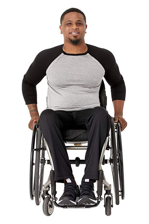 A man with a beard sitting in a manual wheelchair, wearing a black and gray raglan t-shirt, black pants, and black sneakers, photographed against a solid white background.