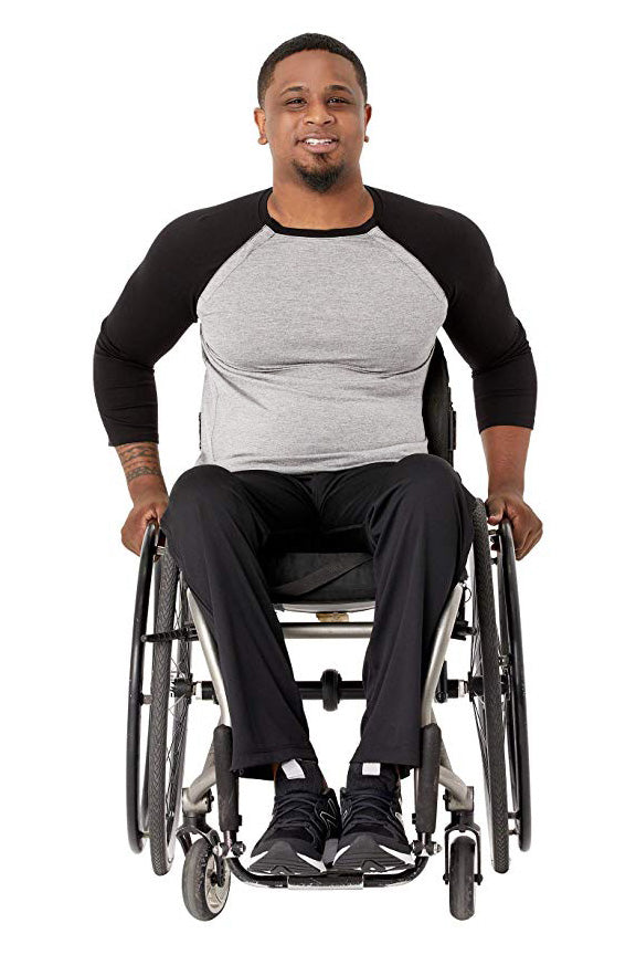 A man with a beard sitting in a manual wheelchair, wearing a black and gray raglan t-shirt, black pants, and black sneakers, photographed against a solid white background.