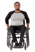 A man with a beard sitting in a manual wheelchair, wearing a black and gray raglan t-shirt, black pants, and black sneakers, photographed against a solid white background.