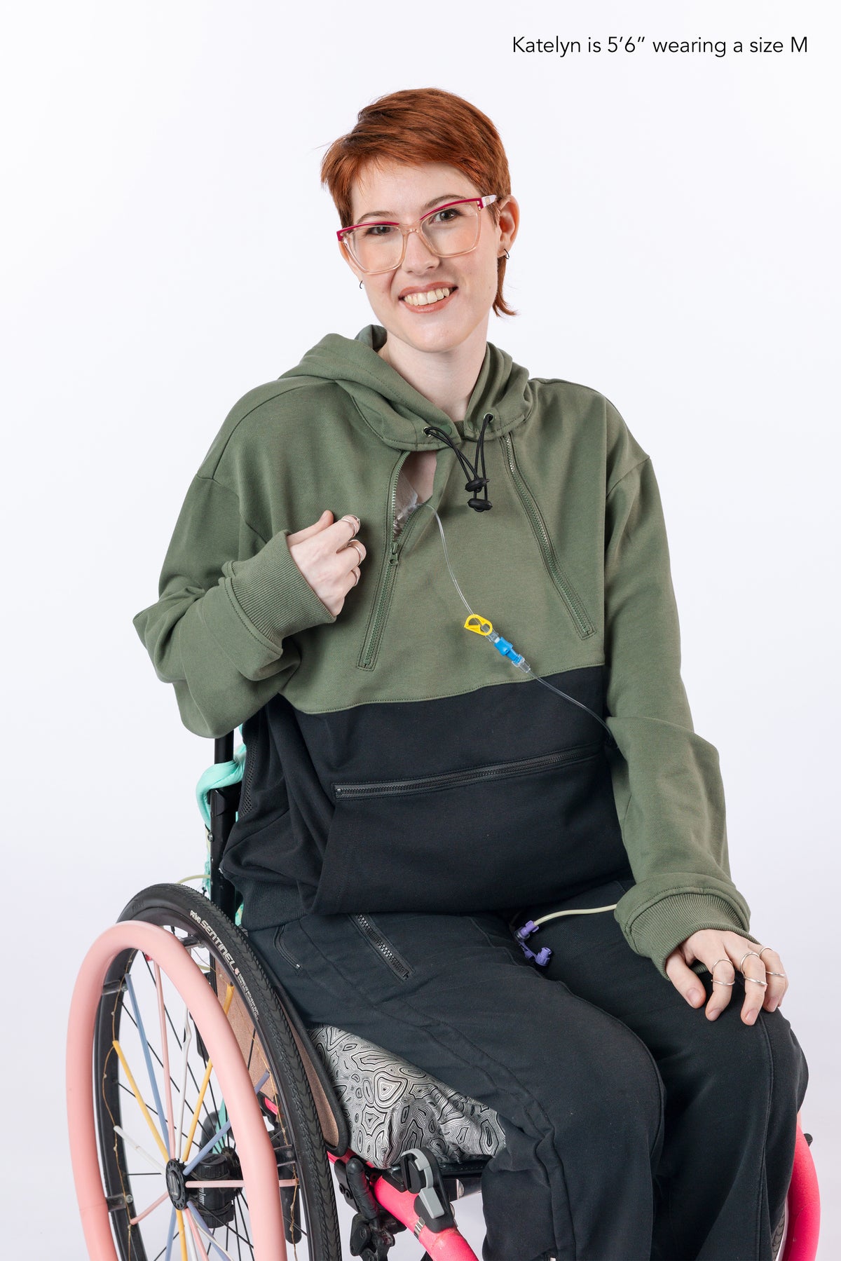 Model wearing Sage Colorblock Hoodie with PICC line coming out of chest zipper. Text reads "Katelyn is 5'6" wearing a size M".