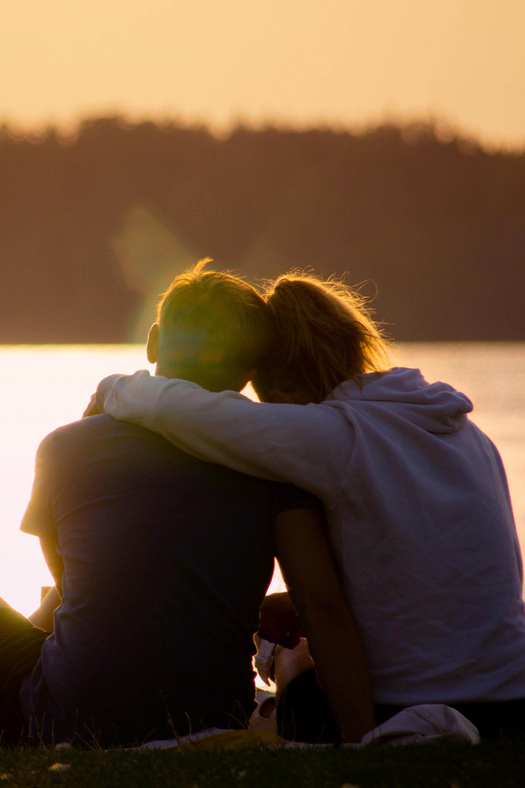 Two people sitting together with a dog at sunset by a body of water.
