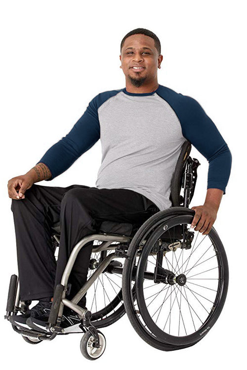 A smiling man in a gray and navy raglan long-sleeve shirt and black pants sits in a manual wheelchair against a plain white background.
