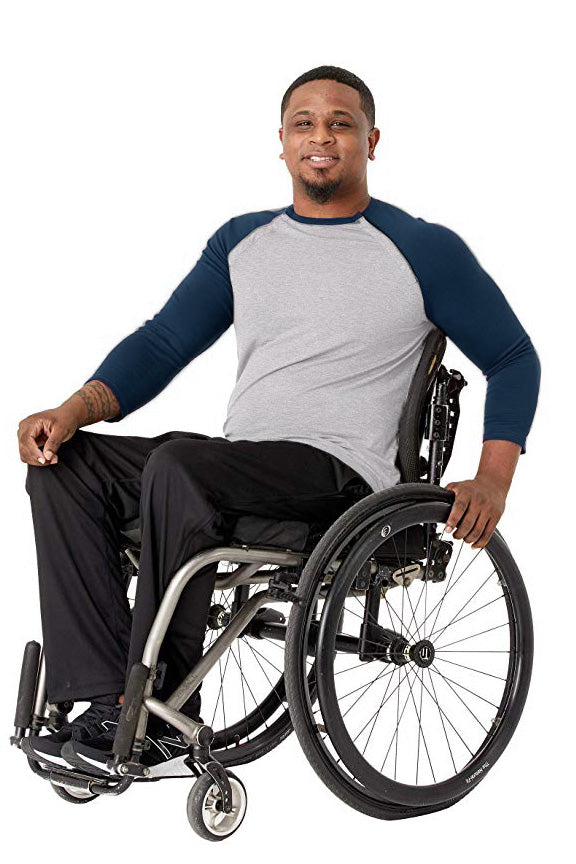 A smiling man in a gray and navy raglan long-sleeve shirt and black pants sits in a manual wheelchair against a plain white background.