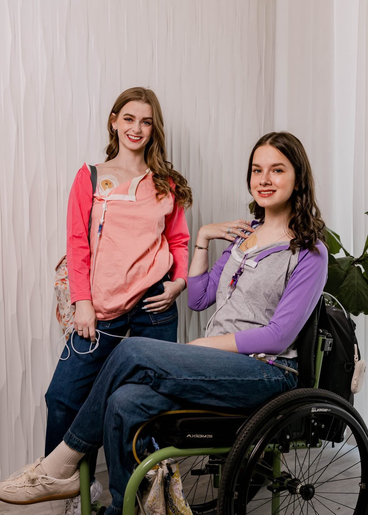 One woman standing in Sunset Raglan Colorblock Access tee, one woman in wheelchair in purple Raglan Access Tee, both showing chest port access