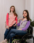 One woman standing in Sunset Raglan Colorblock Access tee, one woman in wheelchair in purple Raglan Access Tee, both showing chest port access