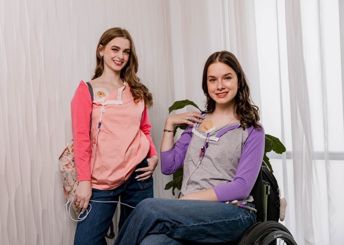 One woman standing in Sunset Raglan Colorblock Access tee, one woman in wheelchair in purple Raglan Access Tee, both showing chest port access