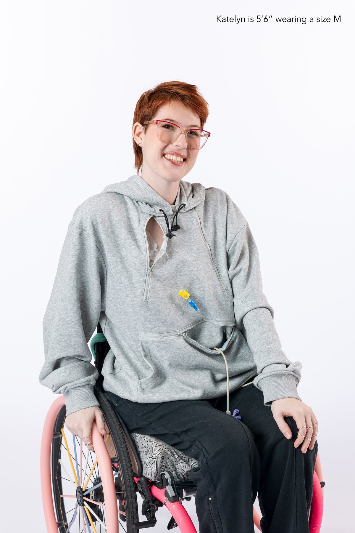 Woman in wheelchair wearing gray zipper access hoodie with open chest zipper with port showing. Text reads "Katelyn is 5'6" wearing a size M".