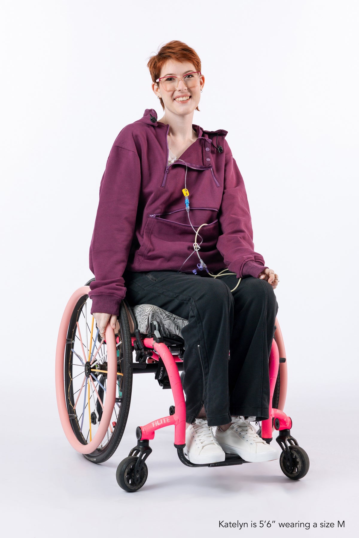 A smiling person with short red hair and glasses sits in a pink manual wheelchair, wearing Plum hoodie with zippers open, black pants, and white sneakers against a plain white studio background. Small text at the bottom reads, 'Katelyn is 5'6" wearing a size M'.