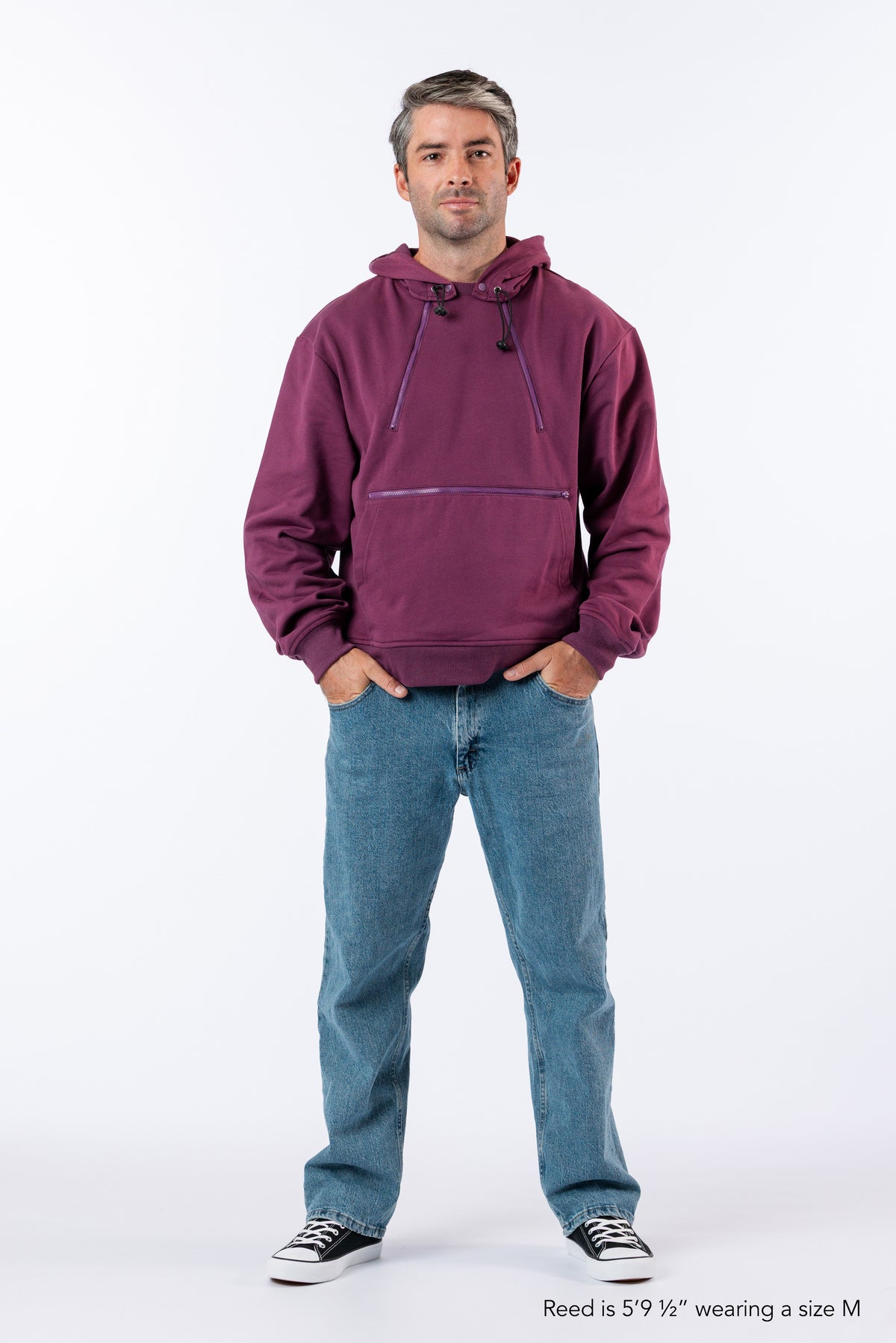 A man modeling a plum zipper access hoodie paired with blue jeans and black-and-white sneakers, against a plain white studio background. Text reads "Reed is 5'9 1/2" wearing a size M".