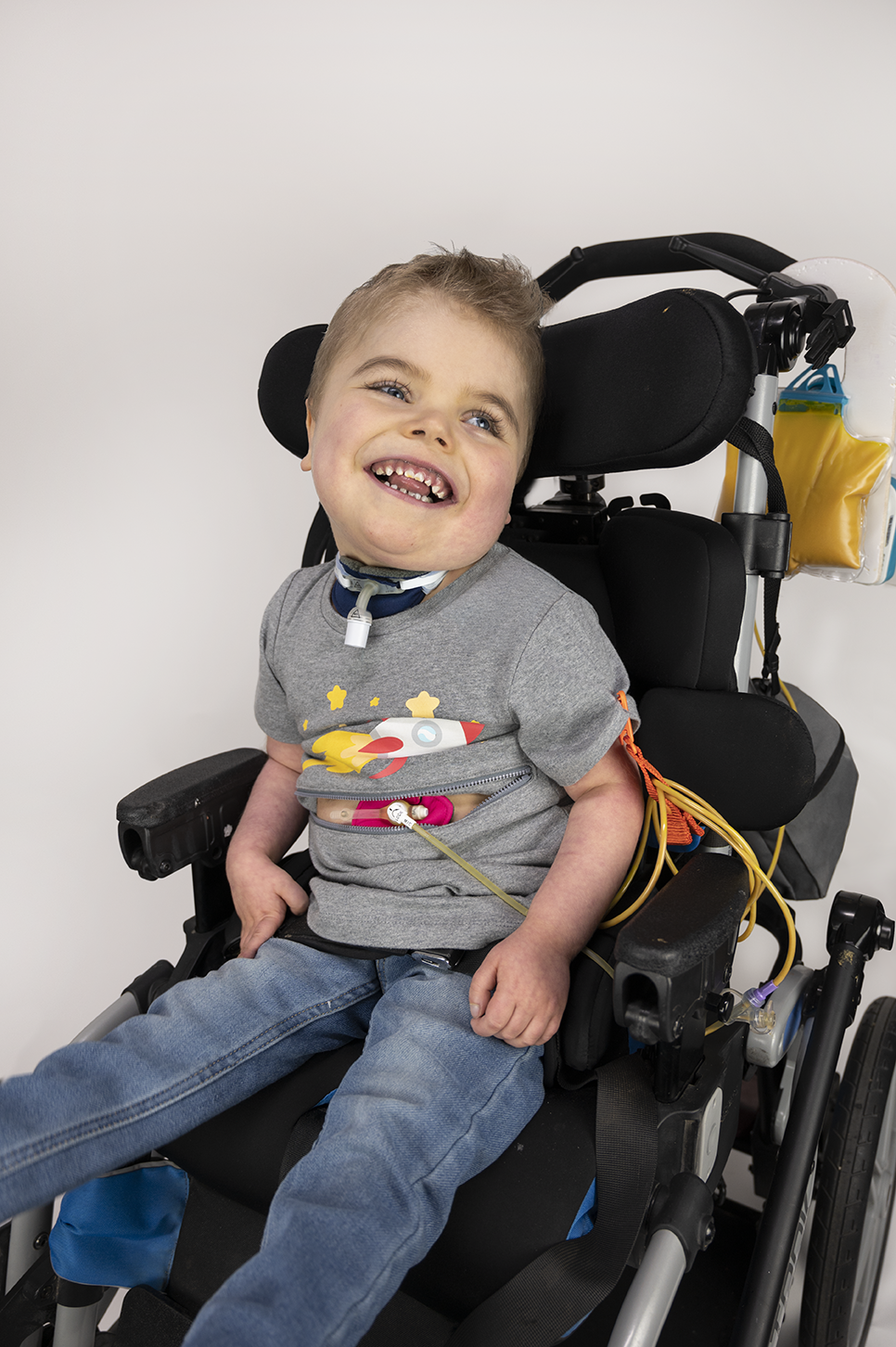 Boy seated in wheelchair wearing Gray Spaceship for Life G-tube zip shirt with zipper for abdominal access open to show g-tube pad