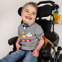 Boy seated in wheelchair wearing Gray Spaceship for Life G-tube zip shirt with zipper for abdominal access open to show g-tube pad